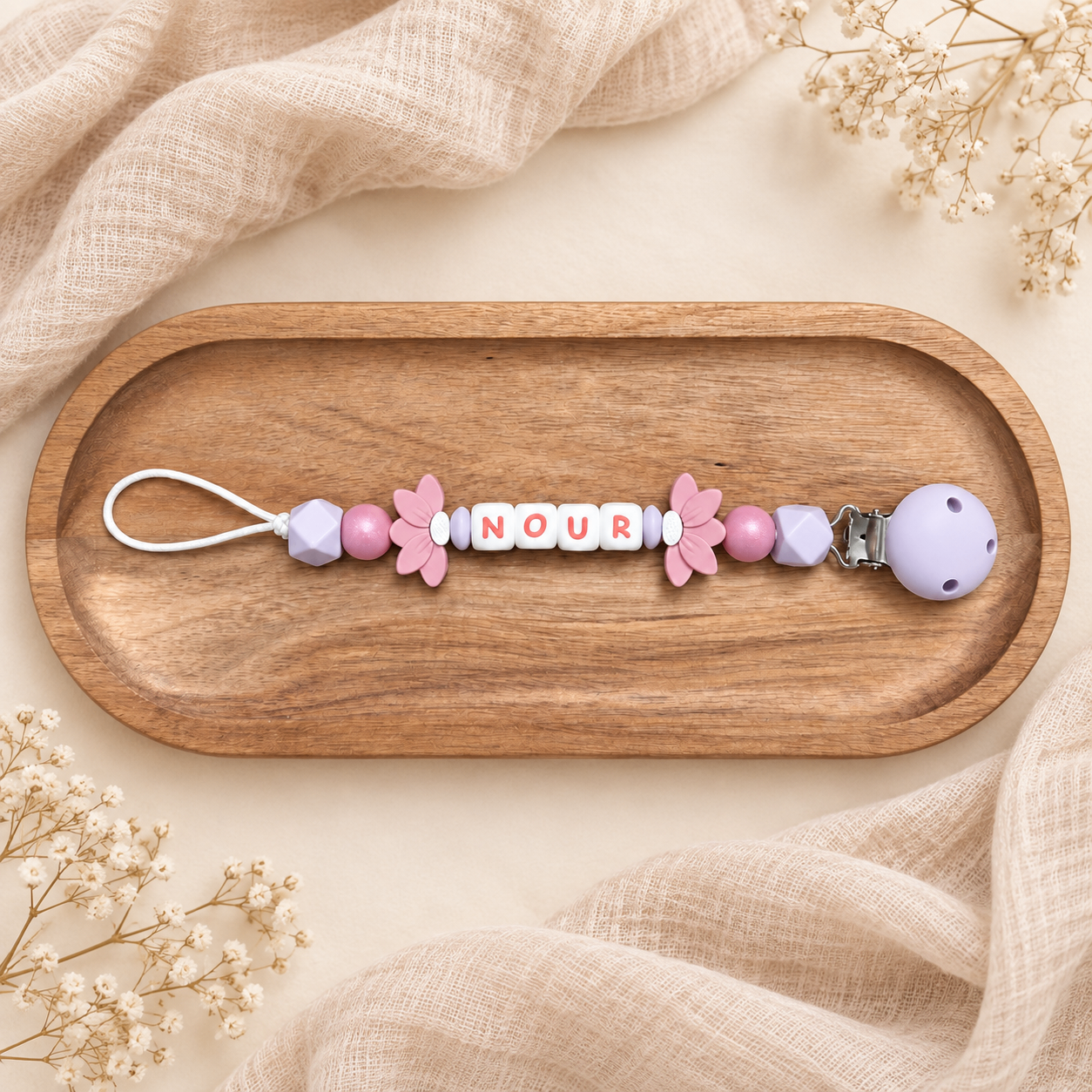 Attache-tétine personnalisée avec perles lilas et roses, fleurs roses et clip mauve, présentée sur un plateau en bois